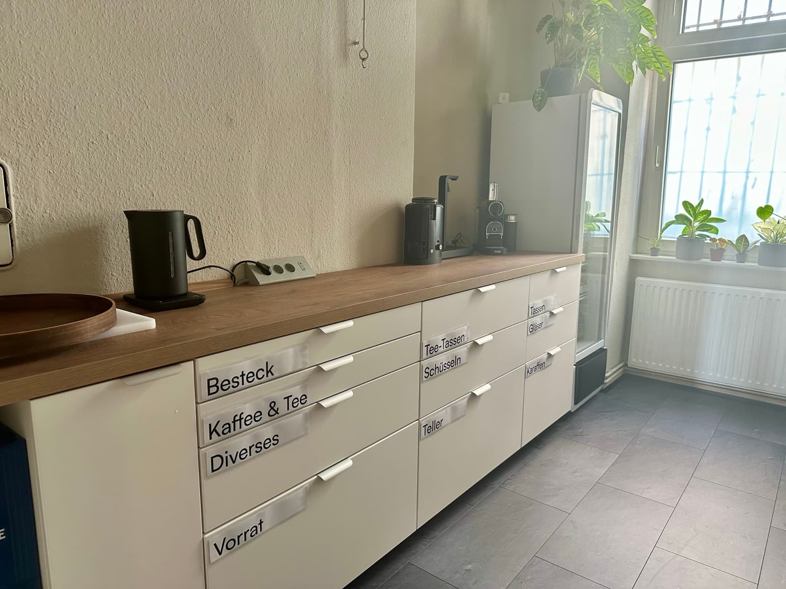 Küche bei Grell37: Küchenzeile mit Eichenplatte und beschrifteten weißen Schubladen, darauf Wasserkocher, Kaffeemühle, Kaffeemaschine und Kapselmaschine, daneben der Getränkekühlschrank am Pflanzenfenster.