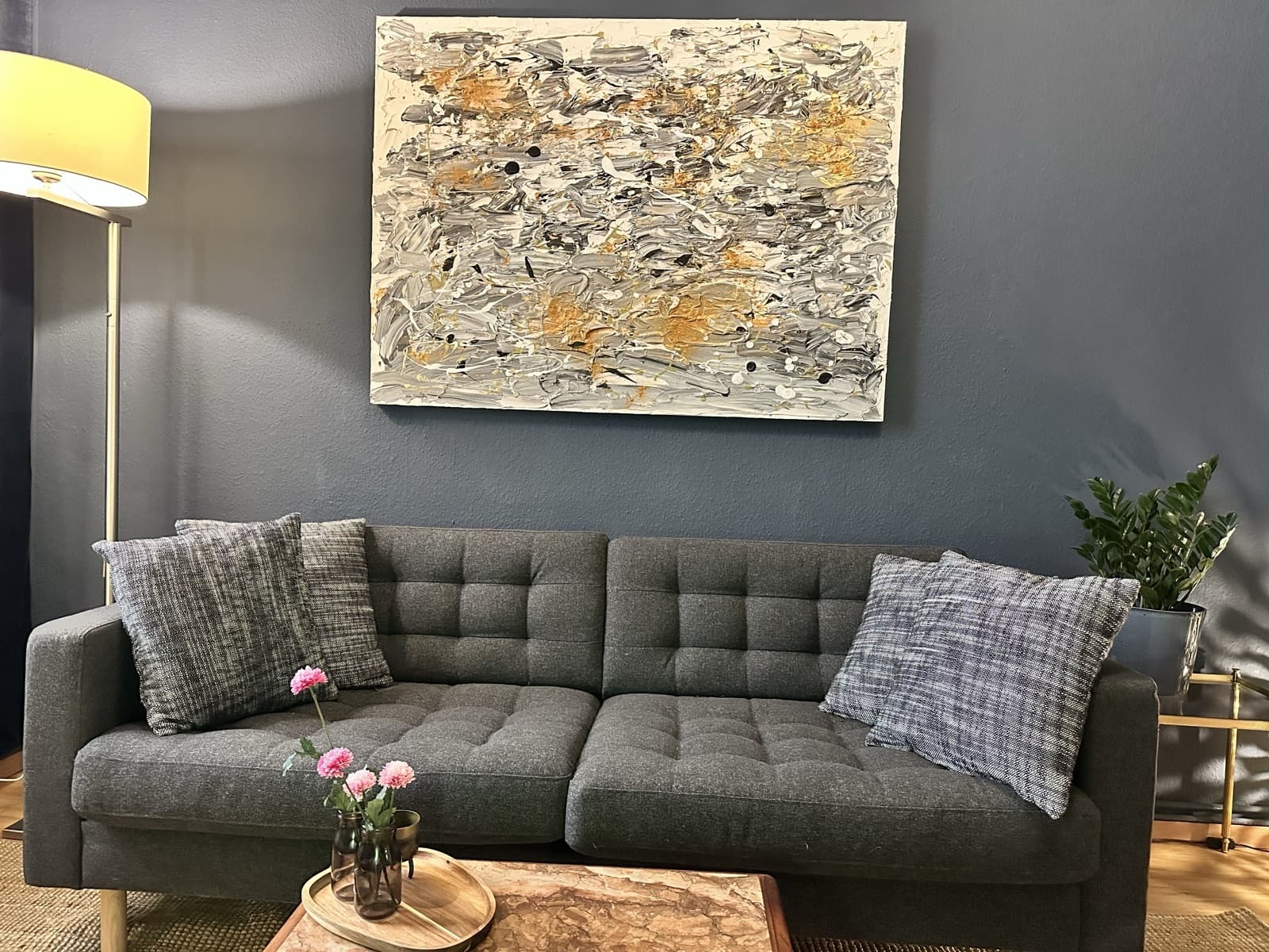 Graue Couch unter einem großen abstrakten Gemälde in Grau und Gold, dunkelblaue Wand und frische Blumen auf dem Couchtisch.