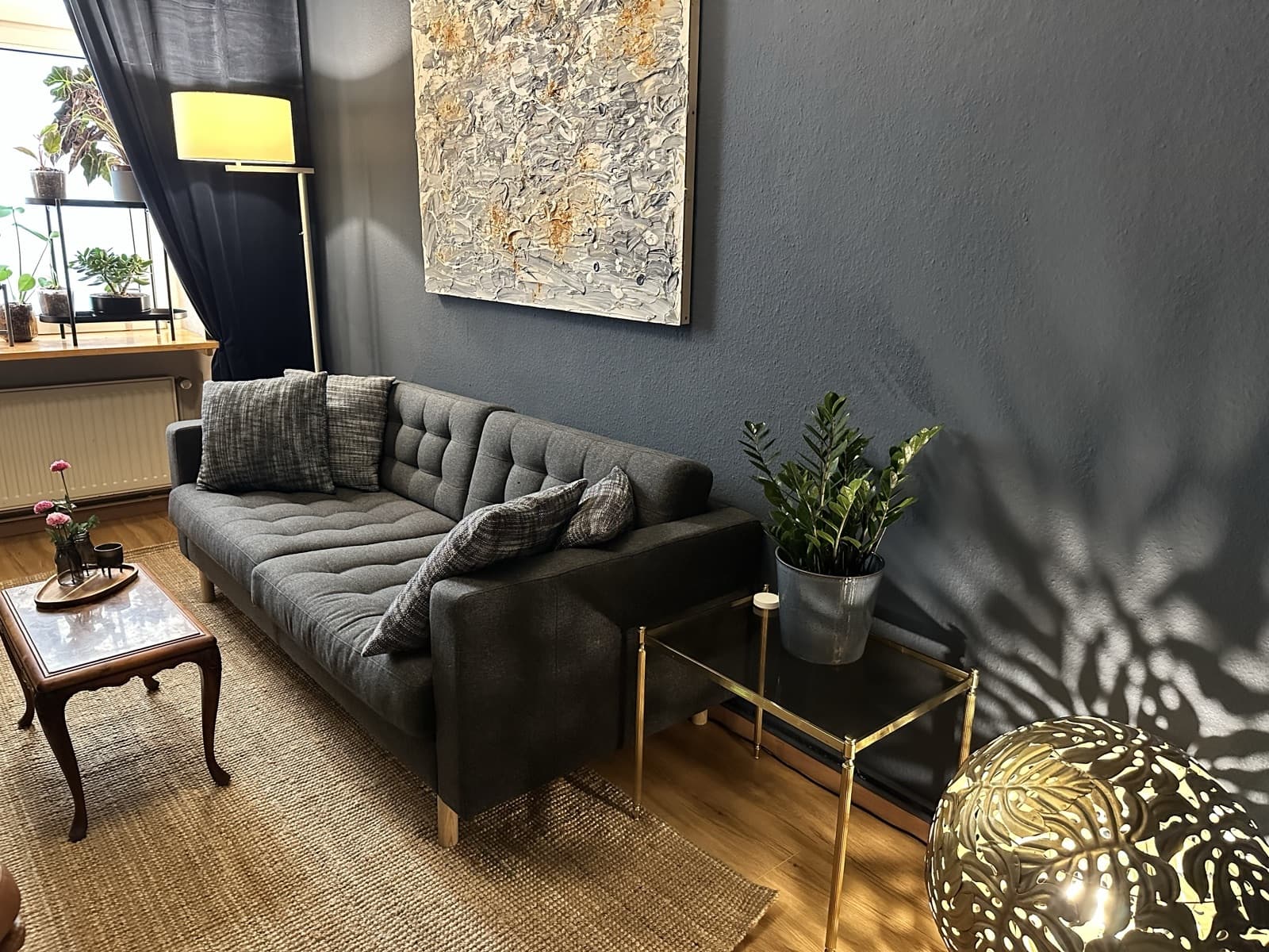 Blick entlang der grauen Couch auf den Messing-Beistelltisch mit Zimmerpflanze, dazu Schattenspiel und Kugelleuchte an der dunkelblauen Wand.