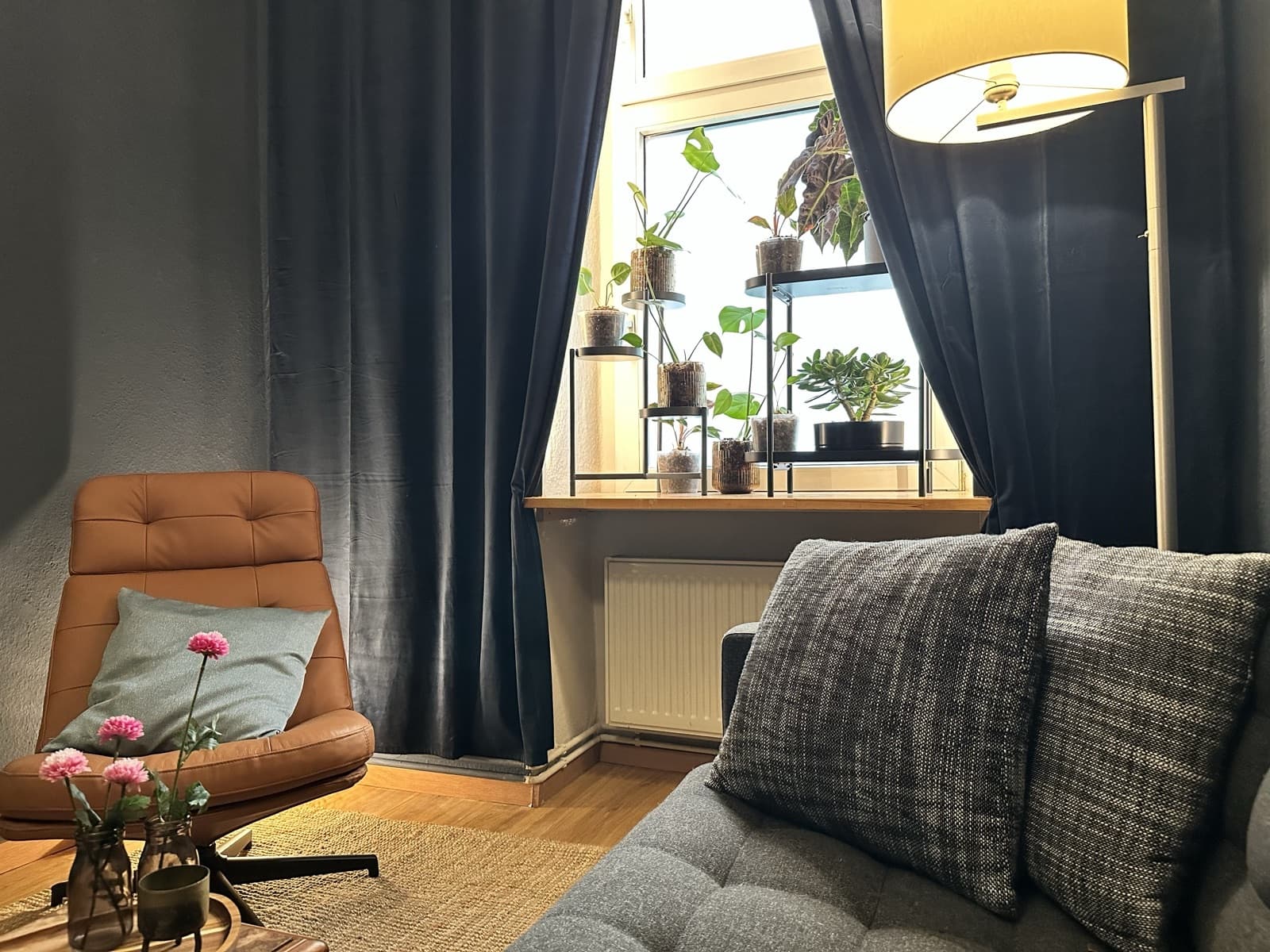 Cognacfarbener Ledersessel mit hellblauem Kissen vor dem Pflanzenfenster der Lounge, dunkelblaue Samtvorhänge und warmes Licht der Stehlampe.