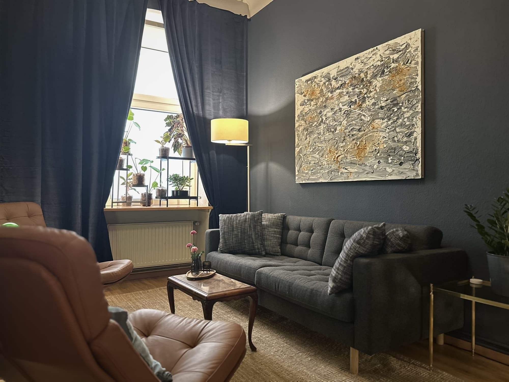 Coaching-Lounge bei Grell37: graue Couch und zwei cognacfarbene Ledersessel um einen kleinen Holztisch, dunkelblaue Wände, Messing-Beistelltische, Pflanzen am Fenster und warmes Licht.