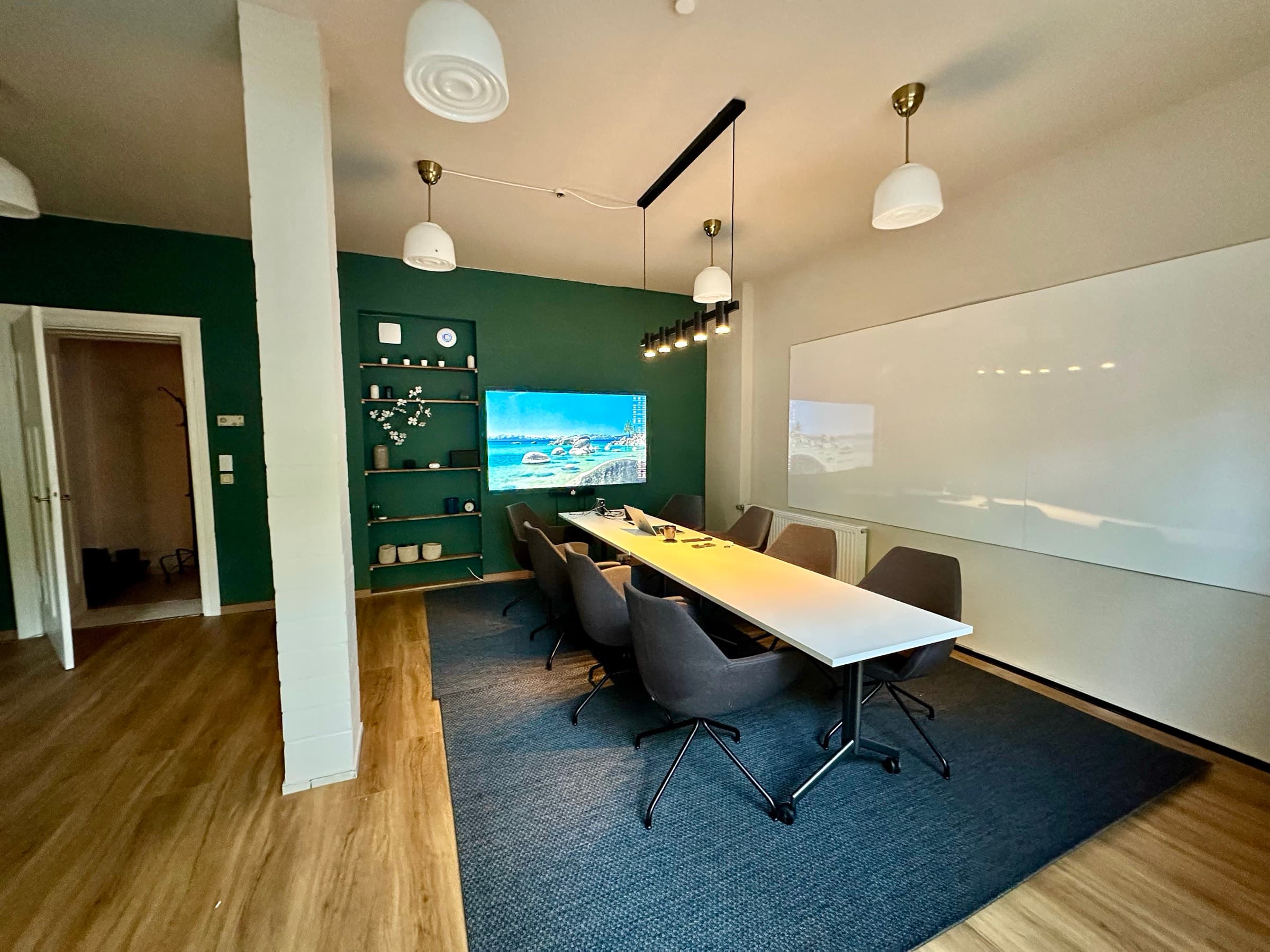 Workshop- und Seminarraum bei Grell37: langer weißer Tisch mit acht grauen Stühlen auf blauem Teppich, 4,5 qm Whiteboard-Wand rechts, 75-Zoll-Bildschirm auf waldgrüner Wand, Messing-Pendelleuchten.