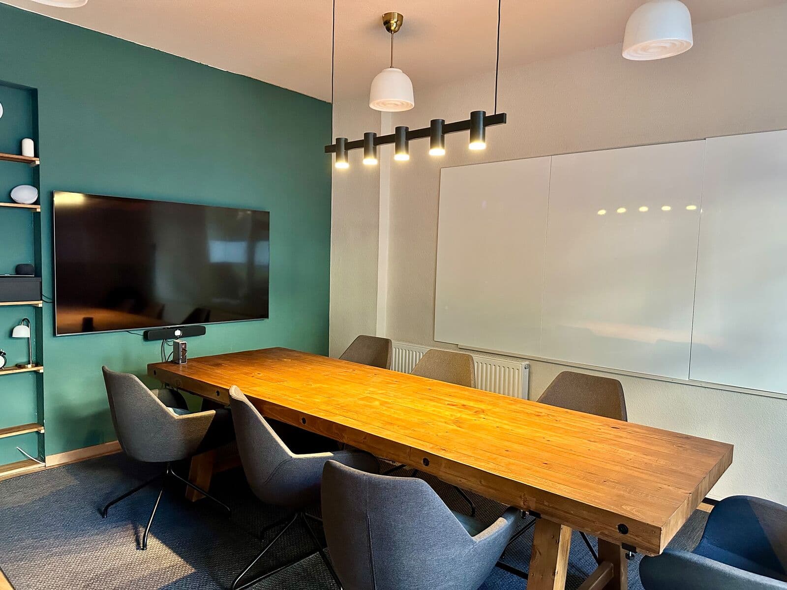 Workshopraum bei Grell37: langer Holztisch mit grauen Drehstühlen, links der 75-Zoll-Bildschirm mit Konferenz-Soundbar auf waldgrüner Wand, rechts die 4,5 qm Whiteboard-Wand, darüber Pendelleuchten und Spots.